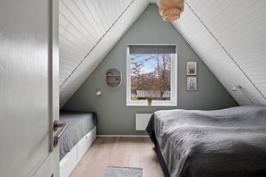 Een slaapkamer met een schuine plafond, een raam met uitzicht op bomen, een bed met een grijze plaid en een klein plankje.