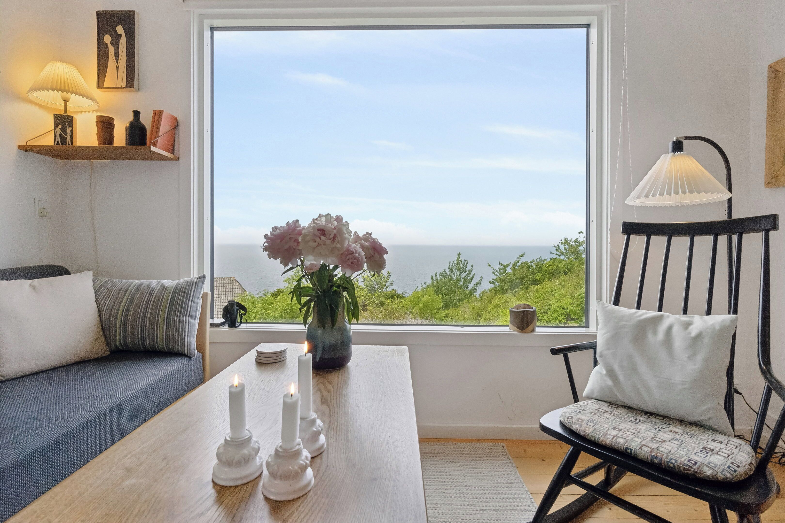 A room with a view of the sea, a wooden table with candles, a black rocking chair, and a sofa.