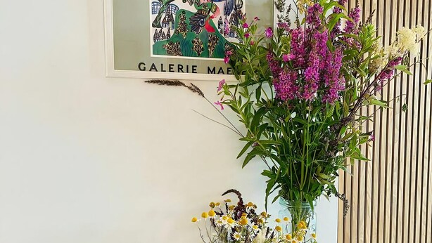 Un mur orné d’un tableau encadré, un pot de fleurs, un vase rempli de fleurs et un saladier de fruits.