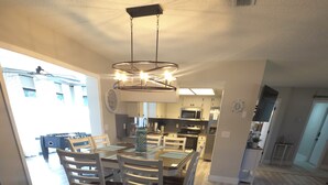 Un vaste espace dédié à la salle à manger, avec une table et des chaises au centre, une cuisine moderne en arrière-plan et un ventilateur de plafond bien visible.