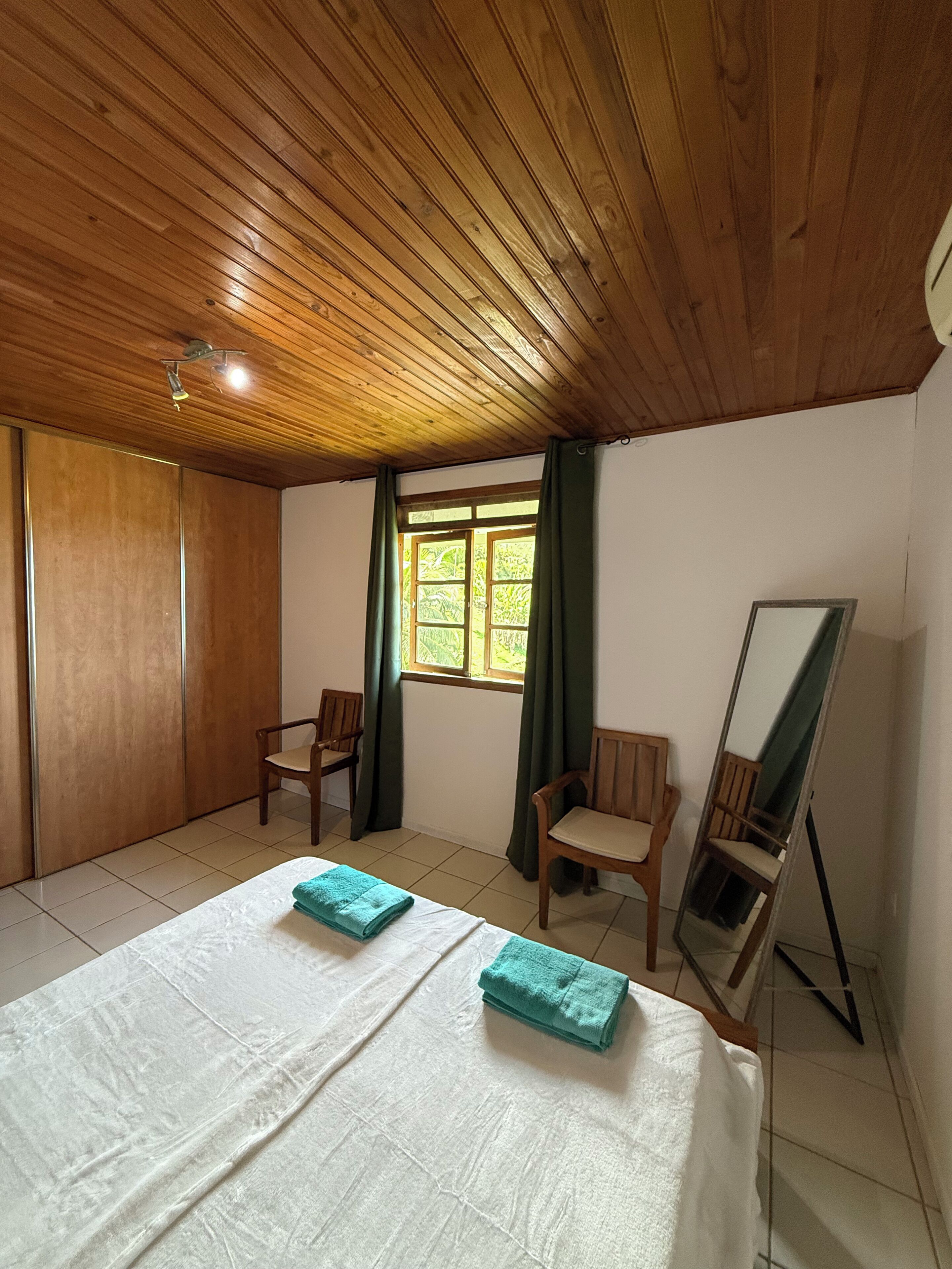 A room with a wooden ceiling, a bed with white bedding and two folded towels, two wooden chairs, a window with green curtains, and a mirror leaning against the wall.