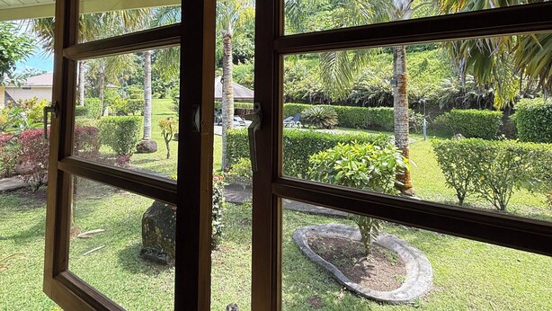 A view through open windows of a well-maintained garden with various plants and a circular flower bed. - Luna's Home Raiatea (Tumaraa)