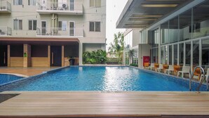 Une piscine avec une terrasse en bois au premier plan, entourée d’un bâtiment moderne doté de balcons et de grandes fenêtres.