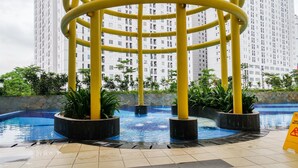 A pool area with a circular water feature surrounded by yellow pillars, set against a backdrop of high-rise residential buildings. - Cozy 2Br At Apartment Bassura City (Jakarta)