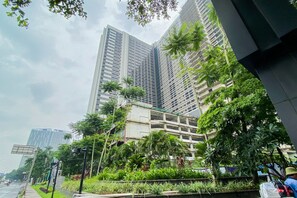 A modern high-rise building with a mix of glass and concrete facades, surrounded by lush greenery and palm trees. - Comfy Dan Warm Studio Soho At Collins Boulevard Apartment (Tangerang)
