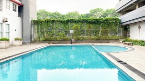 A swimming pool with a tiled deck, surrounded by buildings and greenery. - Cozy Warm Tone Studio At Monroe Tower Apartment (Cikarang)