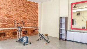 A room with a brick wall, a ceiling fan, and exercise equipment including an elliptical and a recumbent bike. - Trendy And Comfy Studio (No Kitchen) At Celebrity Mansion (Bekasi)