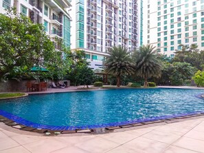 A swimming pool with a tiled deck, surrounded by palm trees and a high-rise building in the background. - Spacious And Lavish 2Br At Woodland Park Residence (Jakarta)