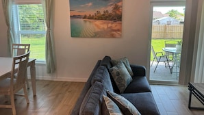A living room with a sofa, a dining table with chairs, a painting of a beach scene, and a sliding glass door leading to an outdoor patio. - Blue Mantle Charlotte Hideaway (Port Charlotte)