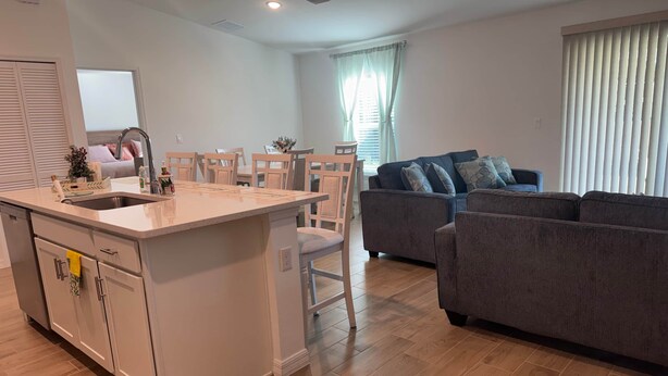 A modern kitchen with a central island, a dining area with chairs, a grey sofa, and a window with blinds. - Blue Mantle Charlotte Hideaway (Port Charlotte)