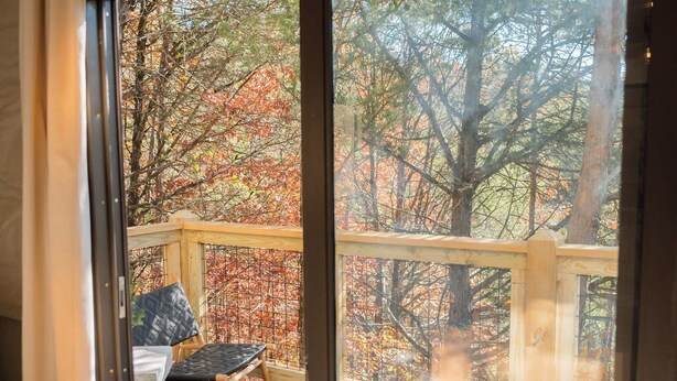 Una terraza de madera con dos sillas negras acolchadas y una mesa, con vistas a un bosque de follaje otoñal.