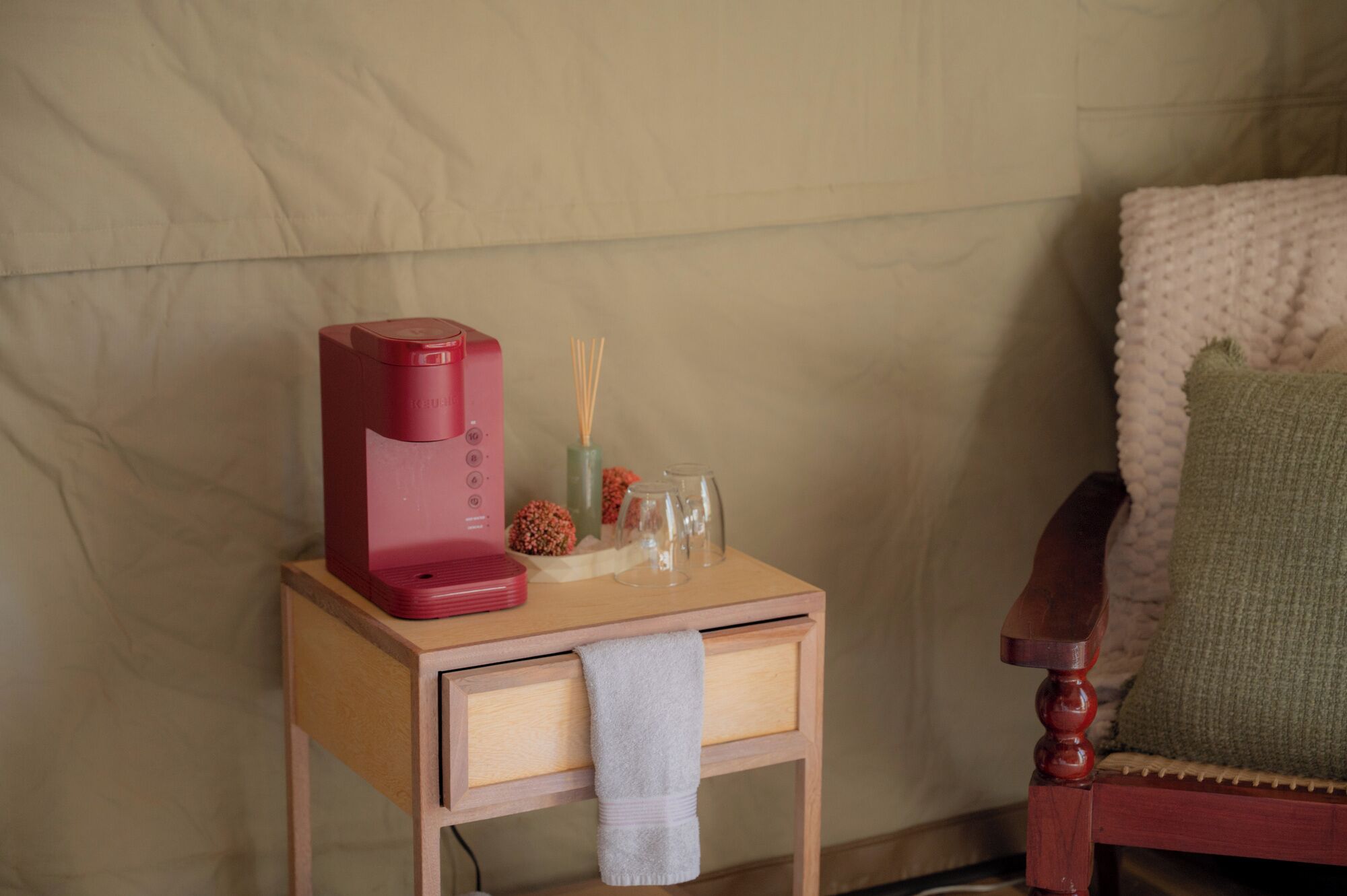 A small wooden bedside table with a red coffee maker, a glass of water, a candle, and a small vase with flowers.