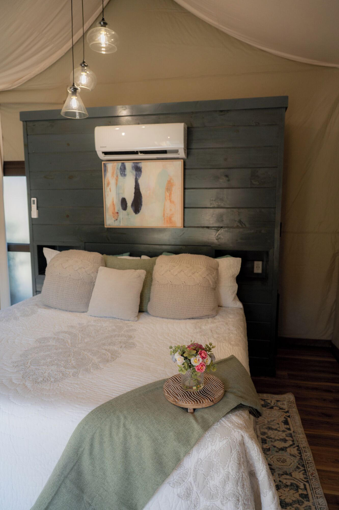 A bedroom with a bed, pillows, a blanket, a vase with flowers, an air conditioner, and a wooden headboard.