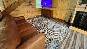 A wooden cabin interior with a brown leather couch, a patterned rug, a television, and a fireplace.