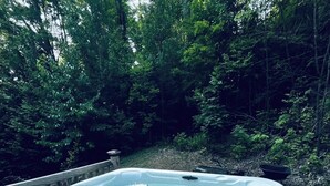 A hot tub in a forested area with a wooden deck and a control panel.