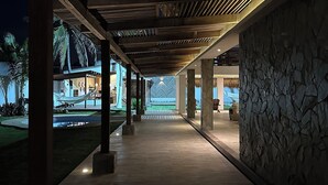 Un espace extérieur abrité, doté d’un plancher en bois, de murs en pierre et offrant une vue sur une piscine et des palmiers en arrière-plan.