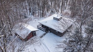 Une cabane moderne dotée d’une grande terrasse et d’un porche abrité, entourée d’une forêt enneigée.