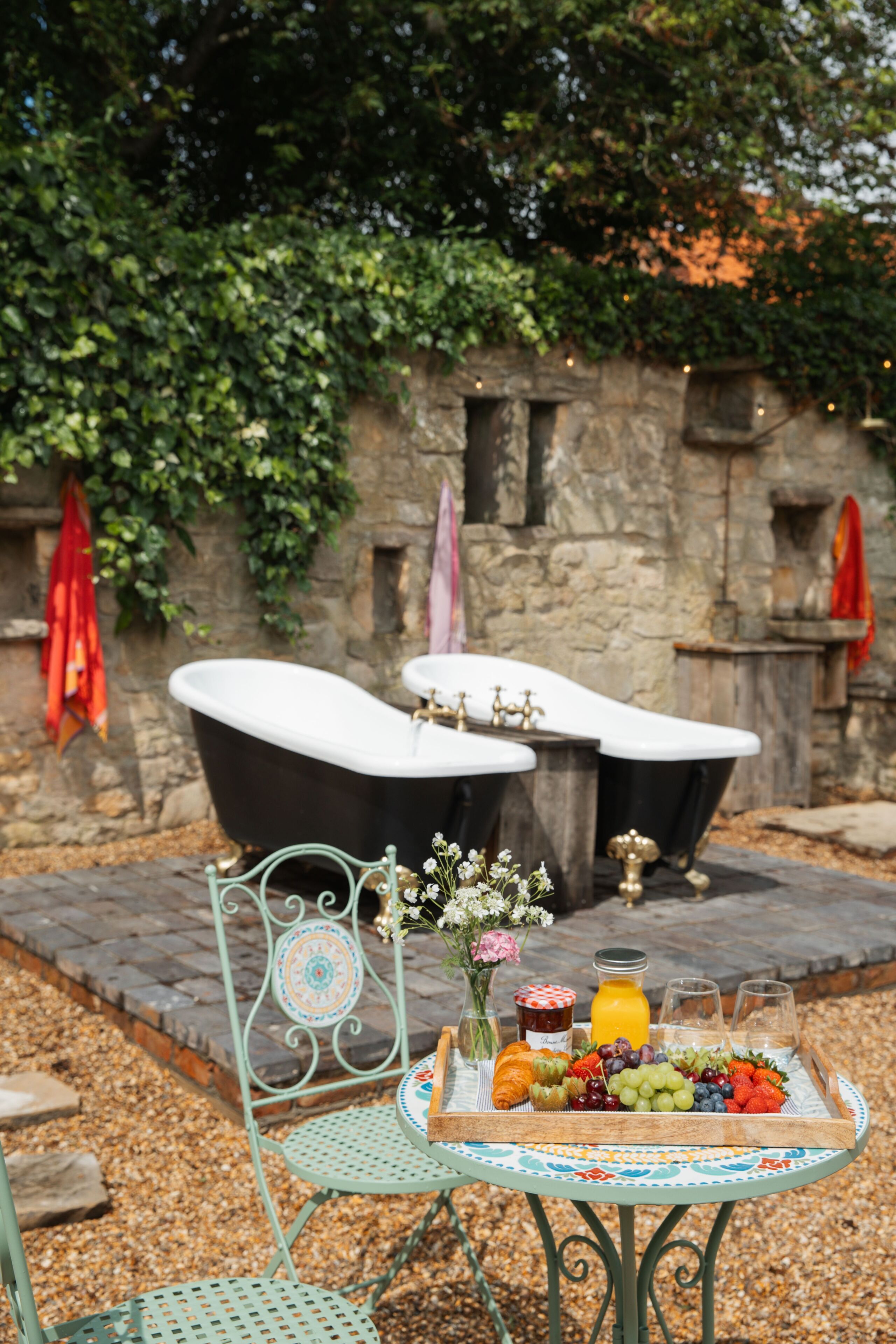 An outdoor seating area with two black tubs, a metal chair, a table with a fruit platter, and a stone wall with greenery.