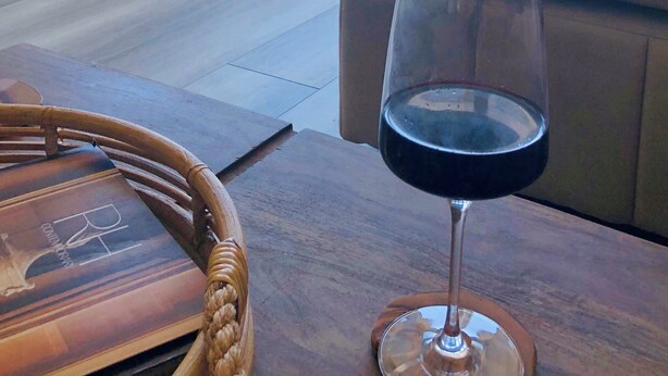 Una habitación con un ventanal grande, una silla blanca, una copa de vino tinto y una mesa de madera.