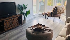 Una sala de estar con televisor, un ventanal grande, ventilador de techo y una planta.