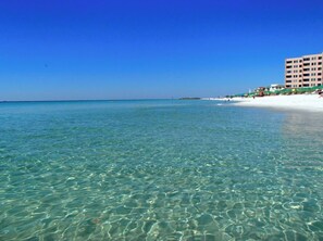 A beach with clear turquoise water, white sandy shore, and a building with multiple balconies. - 🦩Book Me Baby! Islander #615 Aurora in Destin🦩 (Destin)