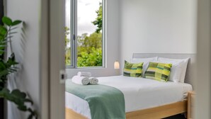 A neatly made bed with a green blanket and two pillows, a window with a view of greenery, and a bedside lamp.