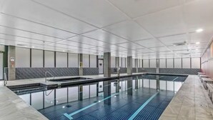 A modern indoor swimming pool with a tiled floor and walls, a clear waterline, and a seating area along one side.