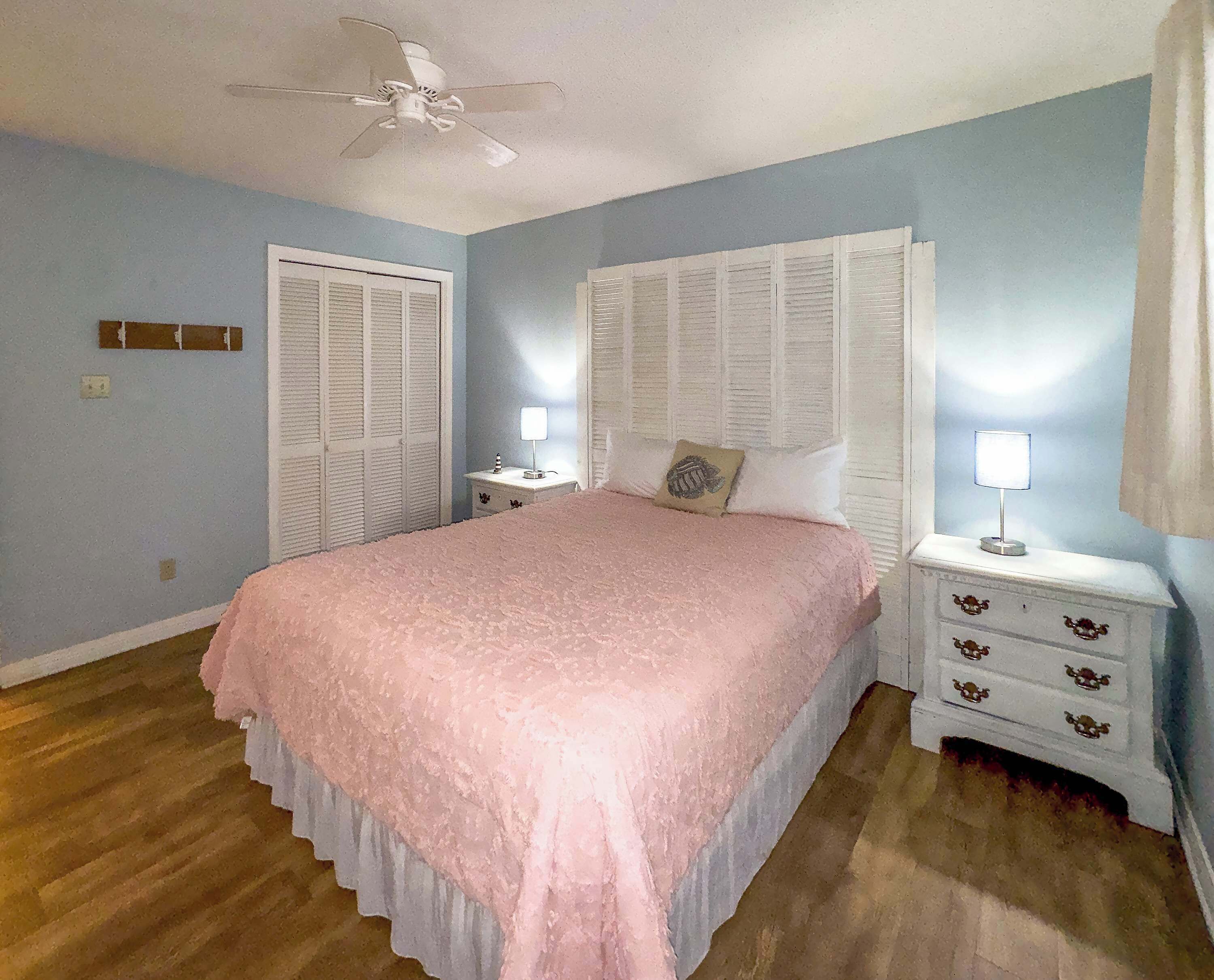 A bedroom with a bed, nightstands, a ceiling fan, and a window with shutters.