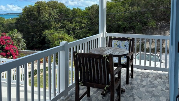 A covered outdoor area with a table and chairs, a white railing, and a view of the ocean and greenery. - Brand New Apartment in George (George Town)