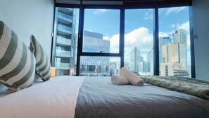 A hotel room with a large bed, pillows, a rolled towel, and a view of tall buildings through large windows. - Cozy & Stylish 1B in the Heart of Brisbane C34 (Brisbane City)