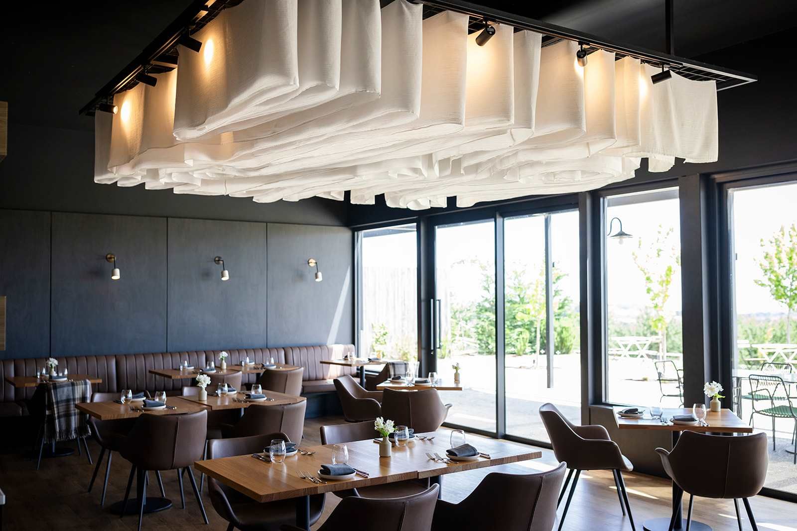 An indoor dining area with tables and chairs, large windows, and a ceiling with hanging fabric.