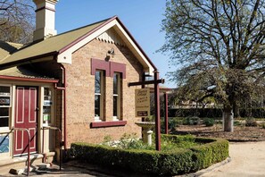 A brick building with a red door and windows, a sign for 'Bastick Cottage,' and a garden area with a hedge and a tree. - Coorabin | Pet-Friendly 4BR near Orange CBD (Orange)