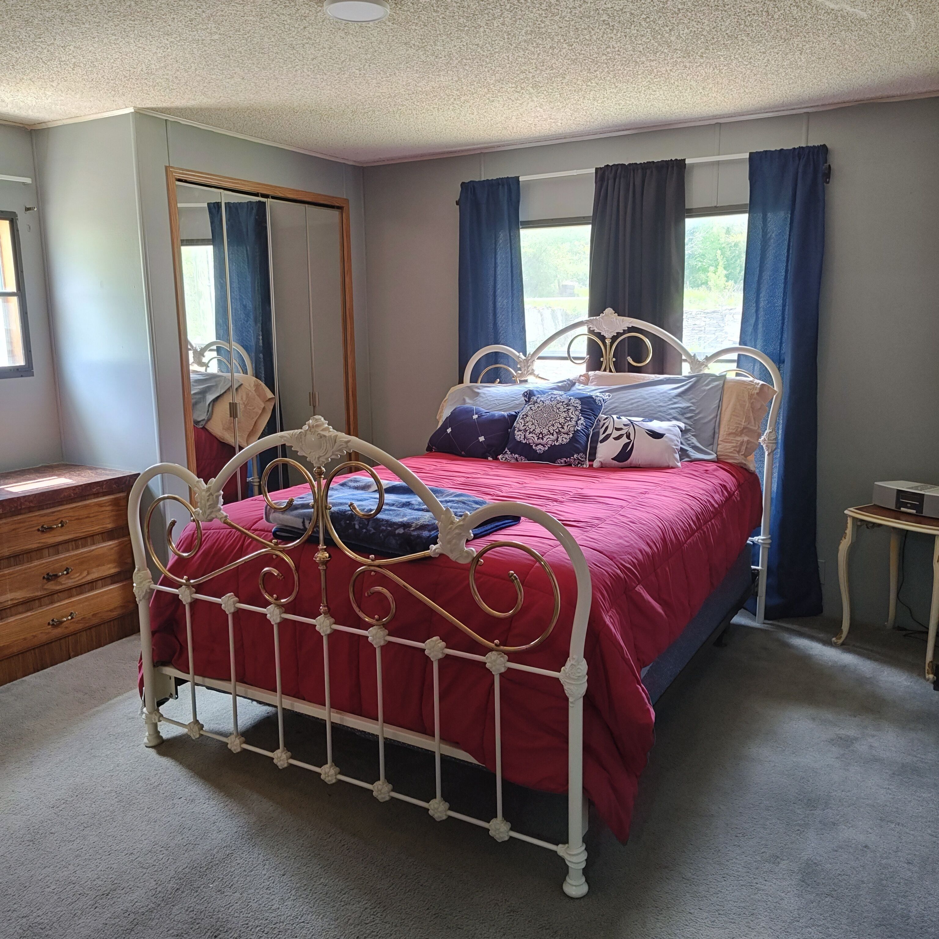 Een slaapkamer met een bed met metalen frame, een houten dressoir, een spiegel en een raam met gordijnen.