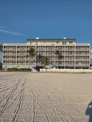 Ein mehrstöckiges Hotel mit einem Balkon an jeder Etage, direkt an einem Strand mit deutlichen Fußabdrücken im Sand.