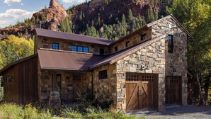 Un edificio rústico de piedra con techo inclinado, grandes puertas de madera para garaje y el paisaje de una montaña al fondo.