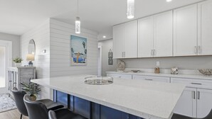 Une cuisine moderne avec des meubles blancs, un grand îlot central doté de tabourets, un éclairage suspendu et un tableau au mur.