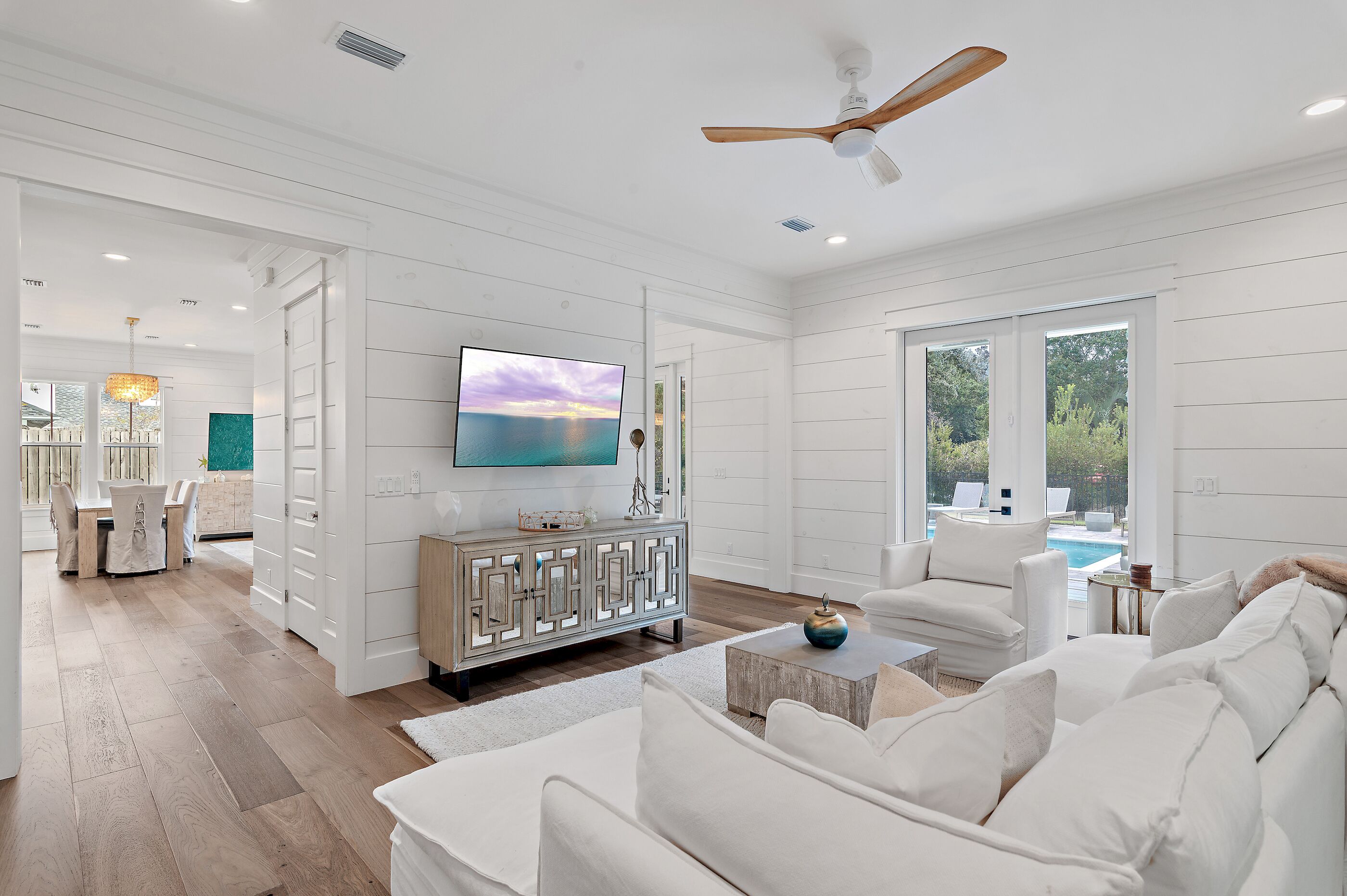 A spacious living room with a white color scheme, a large flat-screen TV, a ceiling fan, and a view of a swimming pool.