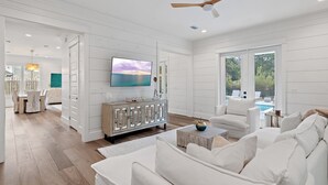 A spacious living room with a white color scheme, a large flat-screen TV, a ceiling fan, and a view of a swimming pool. - 6BR 30A Oasis • Pool, Golf Cart & Bikes (Seagrove Beach)