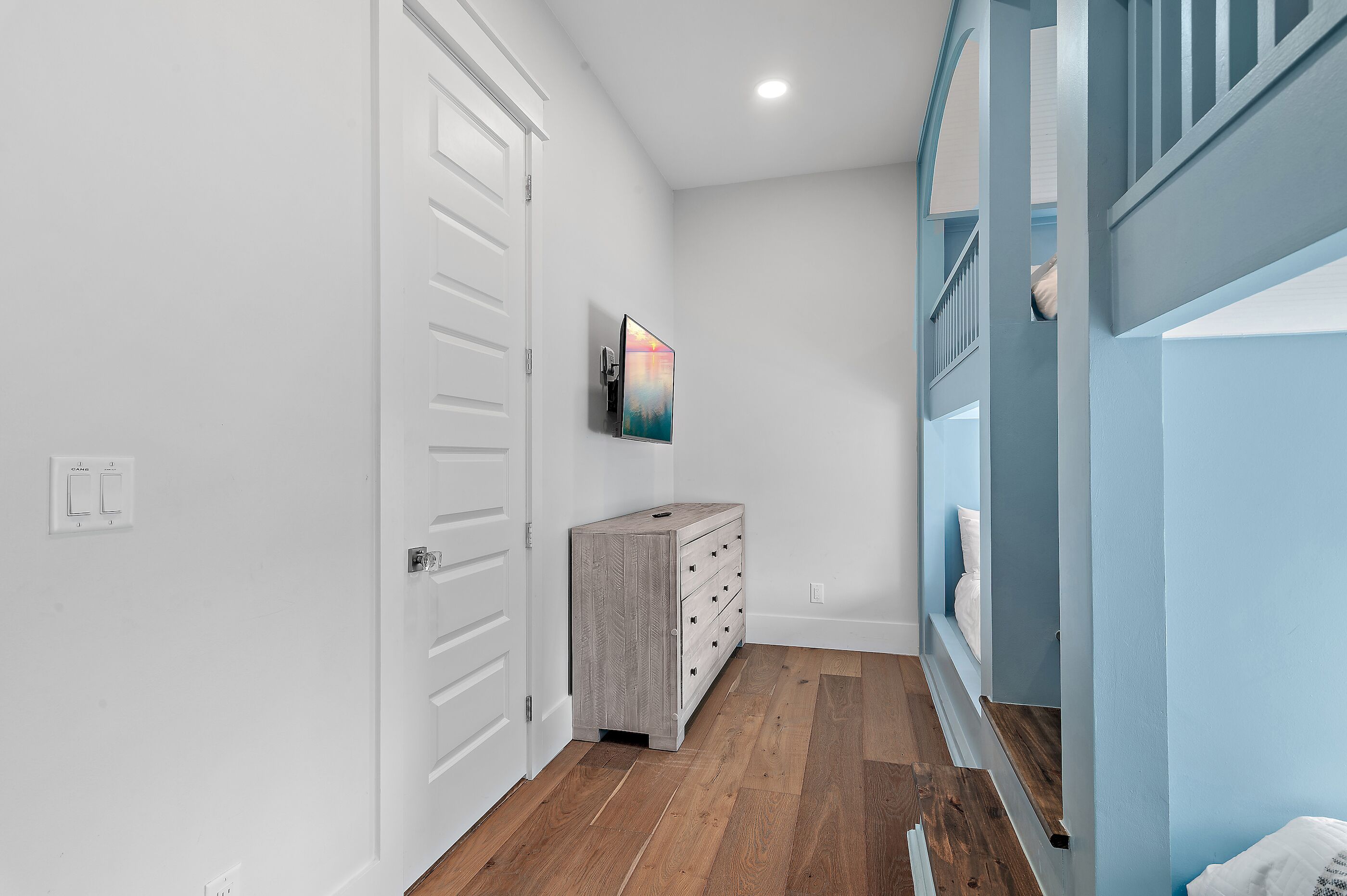 Un couloir avec un plancher en bois, une porte blanche, une commode grise et un tableau accroché au mur.