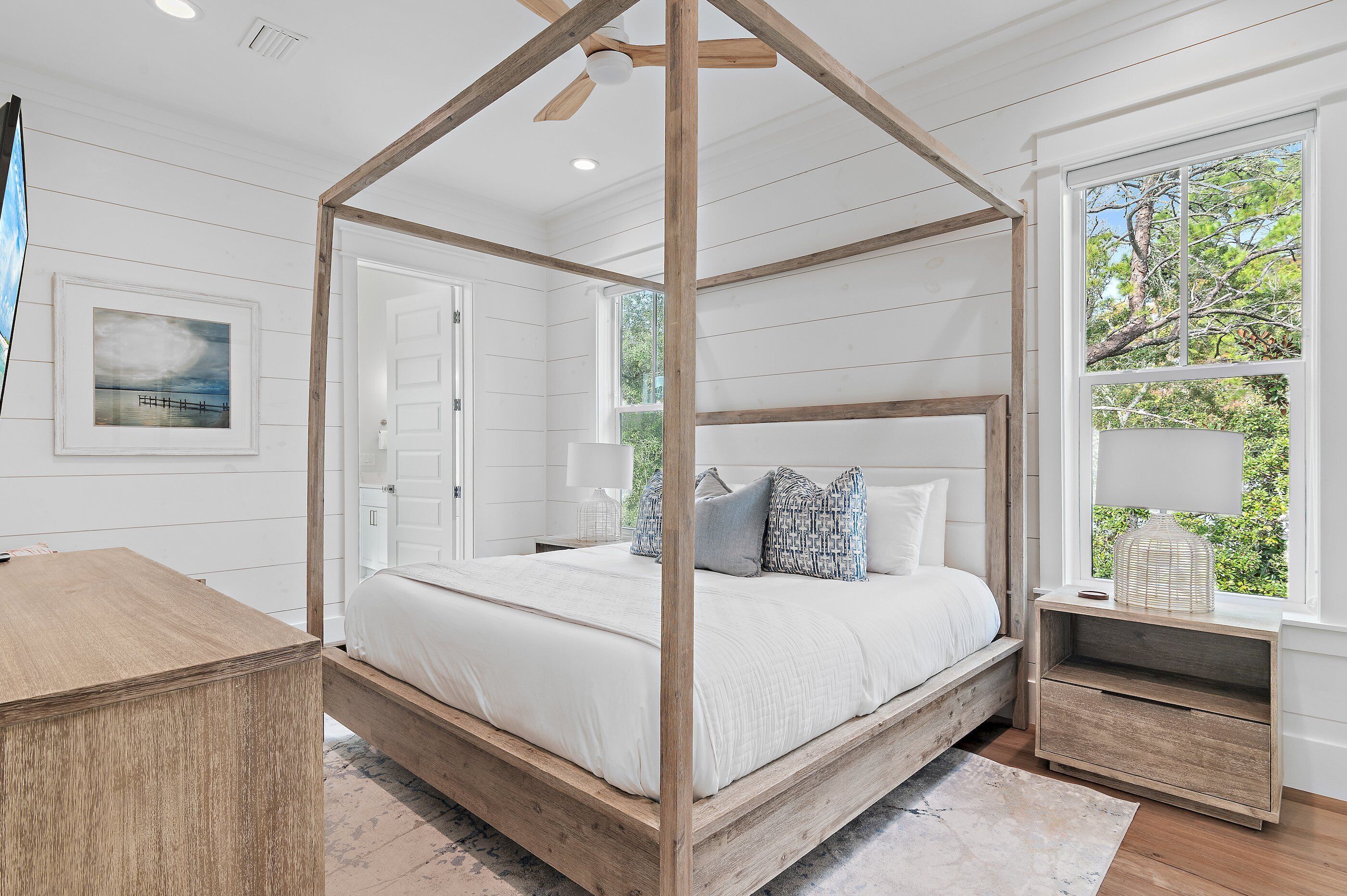 Una cama con dosel en una habitación con paredes de paneles blancos, una mesita de noche de madera y una ventana con vista a árboles.