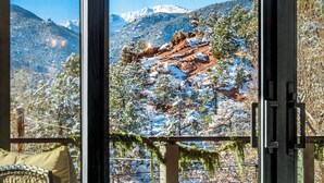 Quarto com grande janela que oferece vista para montanhas nevadas e árvores.
