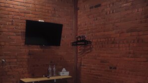 A room with a brick wall, a flat-screen TV mounted above a wooden shelf, a bed with red and white bedding, and a small table with a tissue box and a bottle. - GRHA PAWITRA TROWULAN (Bejijong)