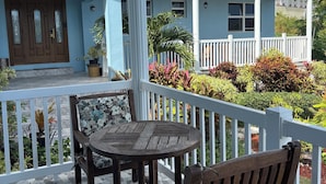 A covered outdoor area with a wooden table and chairs, a white railing, and a view of a two-story building with a balcony.