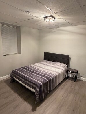 Une chambre à coucher comprenant un lit, une table de chevet, une fenêtre avec des stores et un luminaire au plafond.