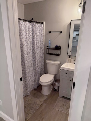 Un baño con inodoro, cortina de ducha, estante con artículos de aseo, espejo y un cubo de basura pequeño.