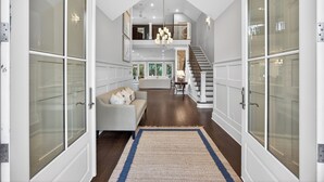 A spacious hallway with a high ceiling, a staircase leading to an upper level, and a cozy seating area with a sofa and cushions. - Expansive Vanderhorst 6Bd Home-- 81 Wax Myrtle (Kiawah Island)