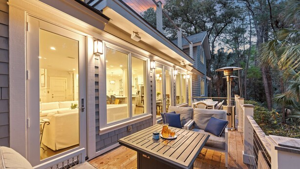 An outdoor patio with a wooden table and seating, large windows, and a view of the interior. - Expansive Vanderhorst 6Bd Home-- 81 Wax Myrtle (Kiawah Island)