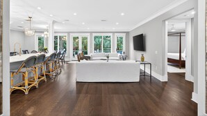 A spacious living room with a large white sofa, a dining area with wicker chairs, and a TV mounted on the wall. - Expansive Vanderhorst 6Bd Home-- 81 Wax Myrtle (Kiawah Island)