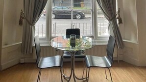Quarto de canto com mesa de vidro redonda, duas cadeiras e vista para a rua através de amplas janelas com cortinas.