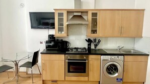 Eine kompakte Küche mit Mikrowelle, Herd, Backofen und Waschmaschine. Ein Glastisch mit Stühlen und ein an der Wand befestigtes Fernsehgerät.